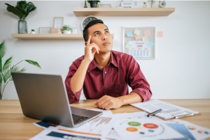 Seorang pria di meja kerja dengan laptop, tumpukan dokumen grafik, dan papan klip kontrak, menopang dagu dan menatap ke atas dengan ekspresi sedang merenung.